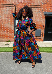 A woman standing with one hand raised wearing a multicolor African print maxi dress with long sleeves, a surplice neckline, and a self tie belt.