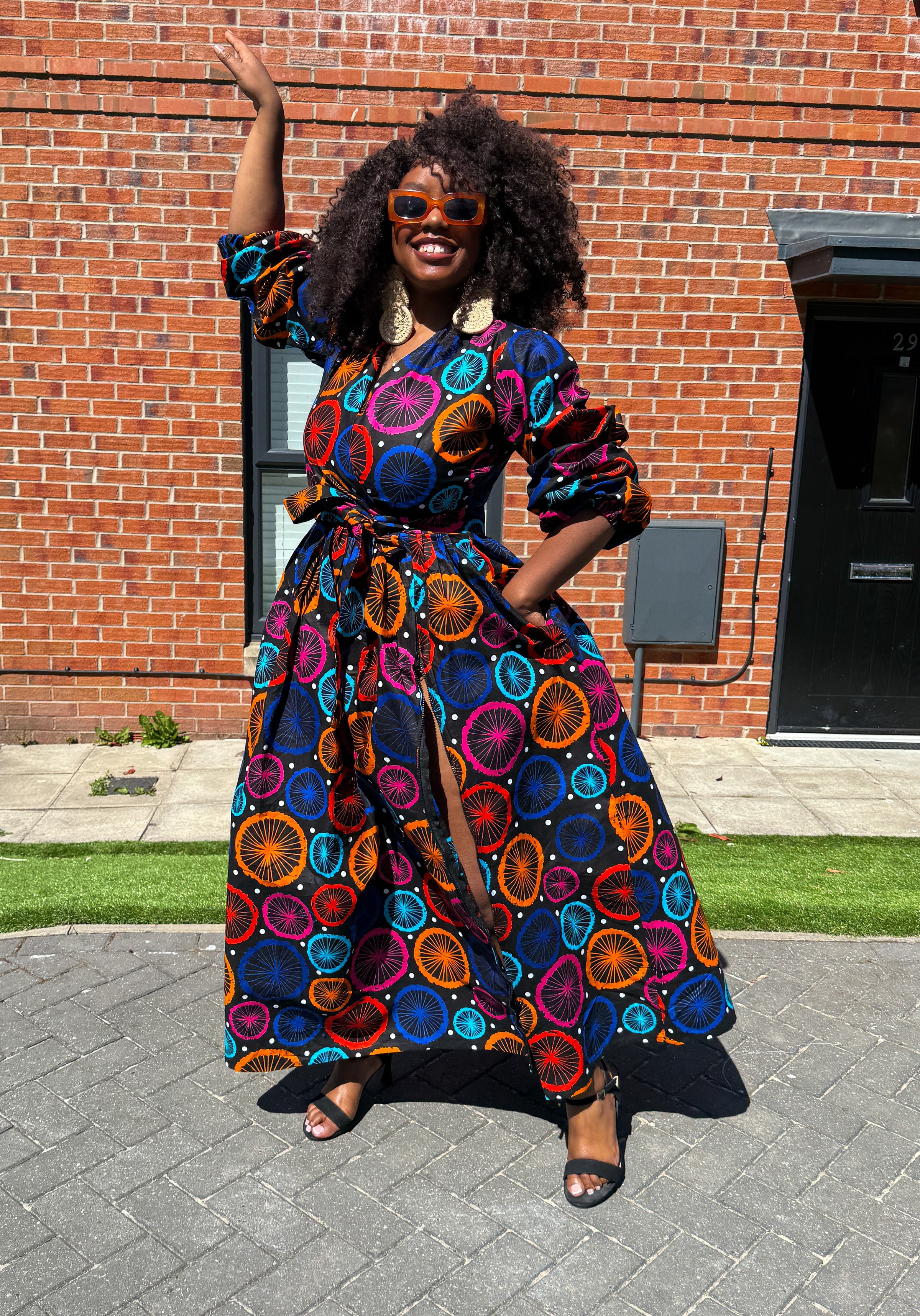 A woman standing with one hand raised wearing a multicolor African print maxi dress with long sleeves, a surplice neckline, and a self tie belt.